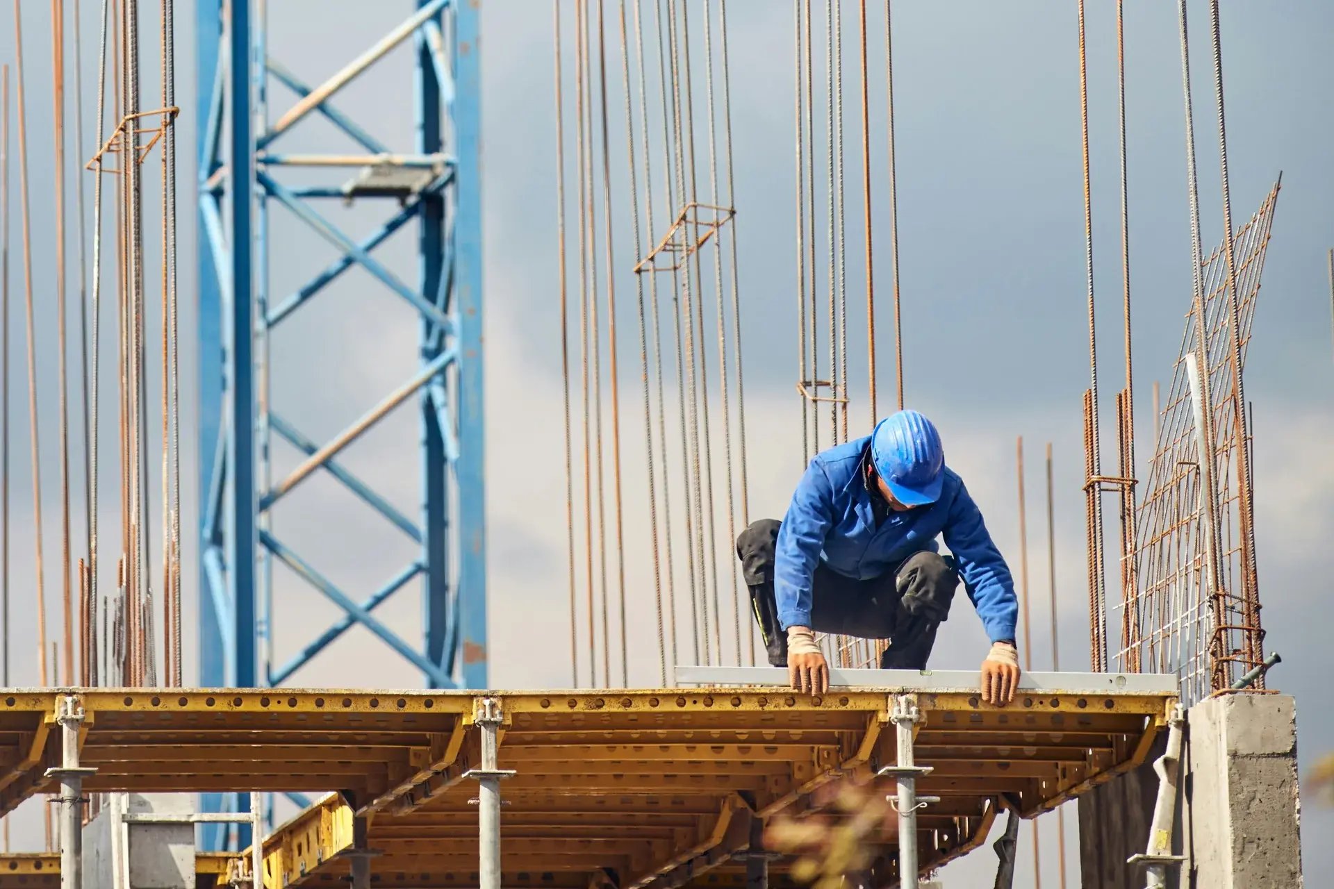 Construction worker in a blue hard hat climbing on rebar formwork at a high-rise building site.