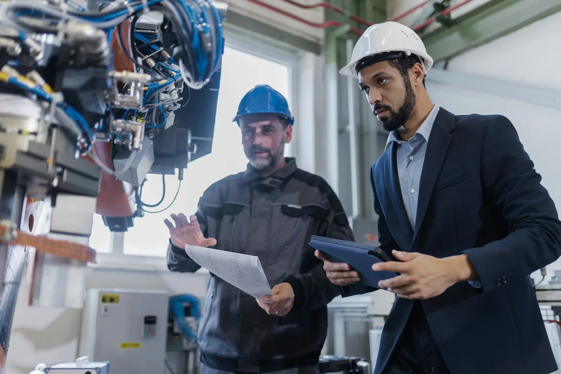 Engineer in a suit and technician in a hard hat reviewing data on a tablet next to industrial robotics equipment.