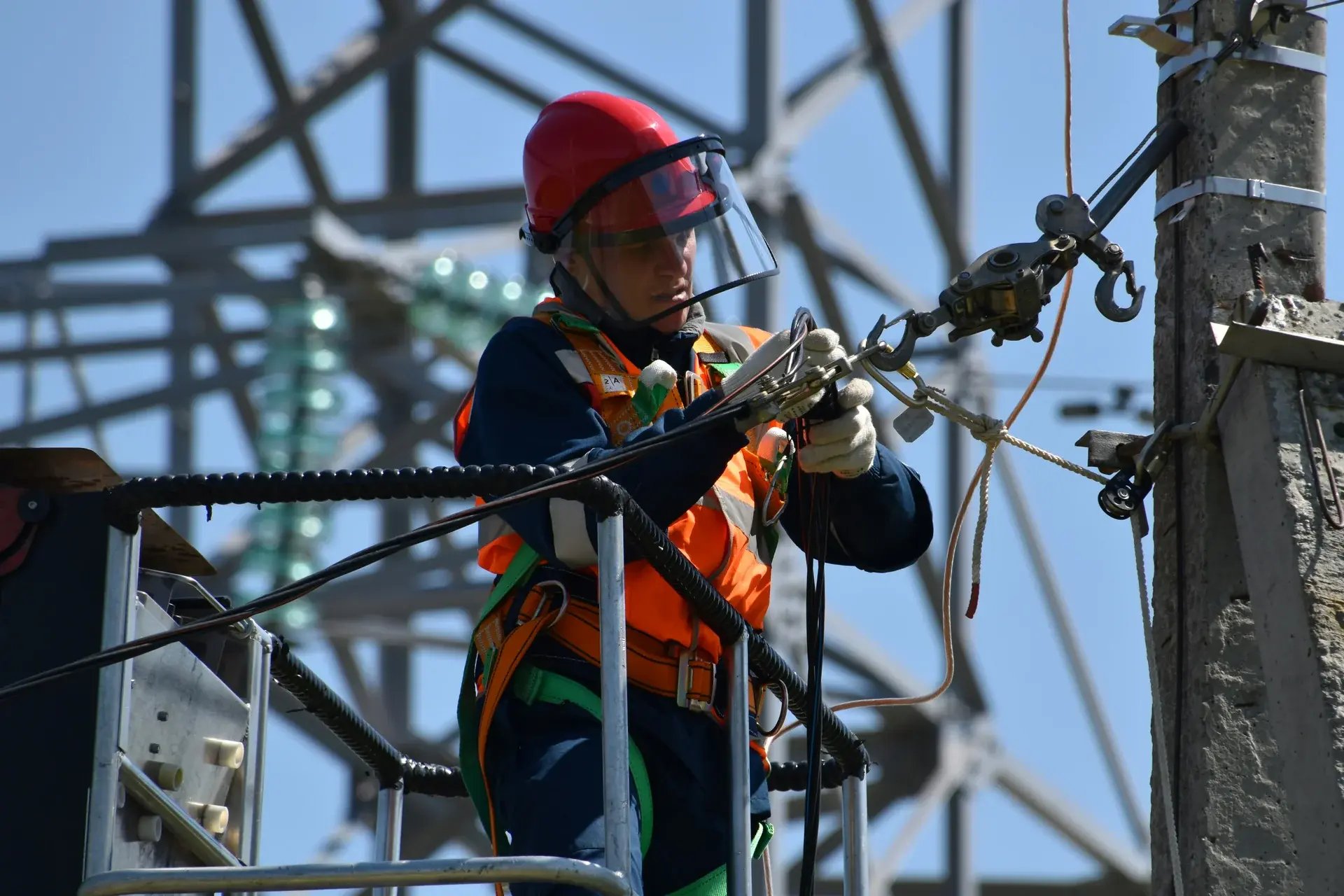 Electrical lineman in safety harness and red helmet working on high-voltage power transmission lines.