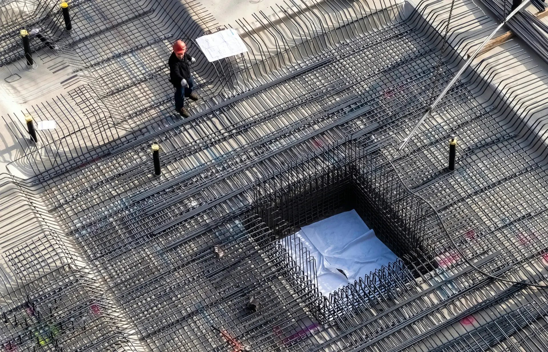 Aerial view of a worker reviewing blueprints on a steel rebar foundation grid at a construction site.