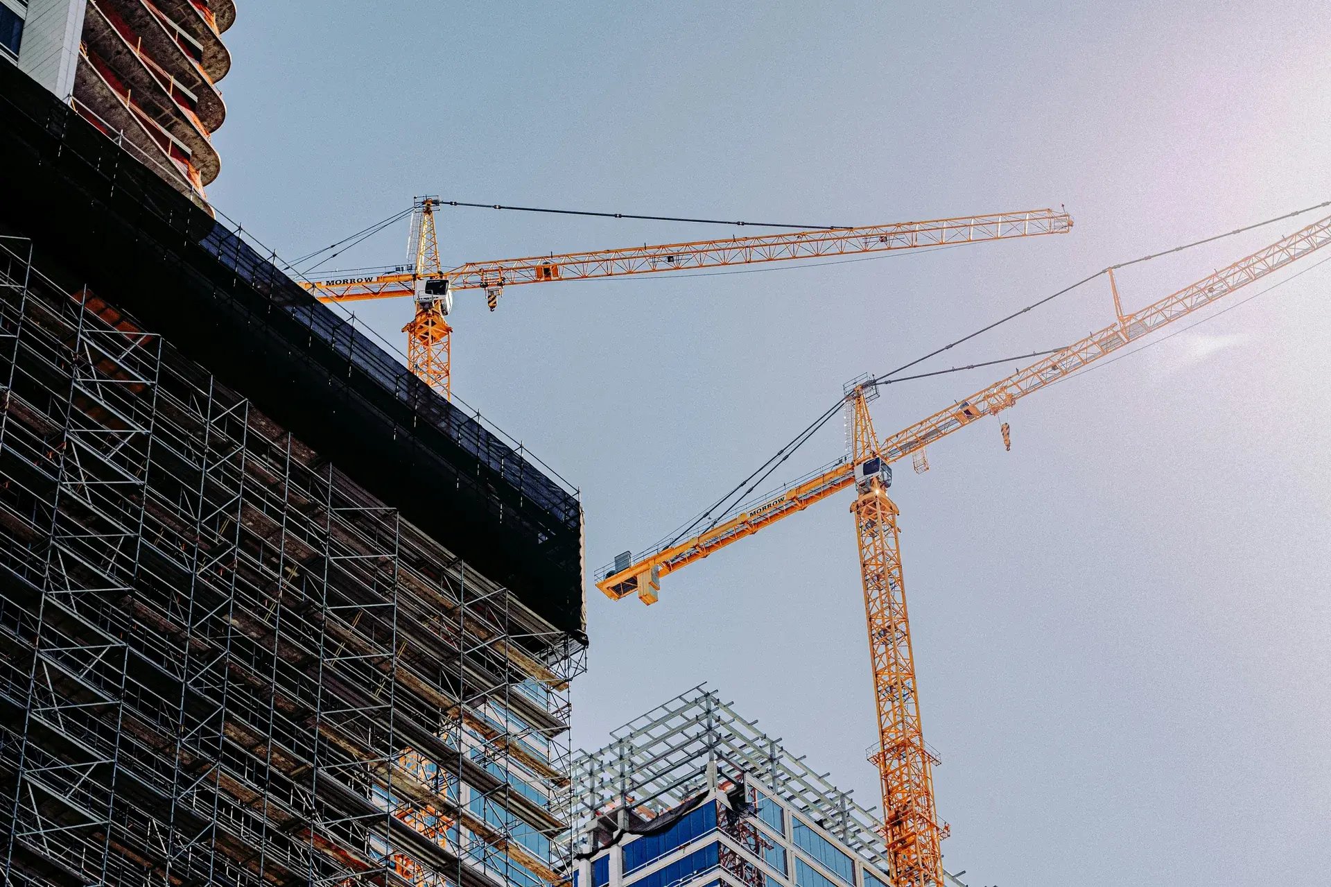 Two tower cranes alongside scaffolding on a high-rise building under construction against a clear urban sky.