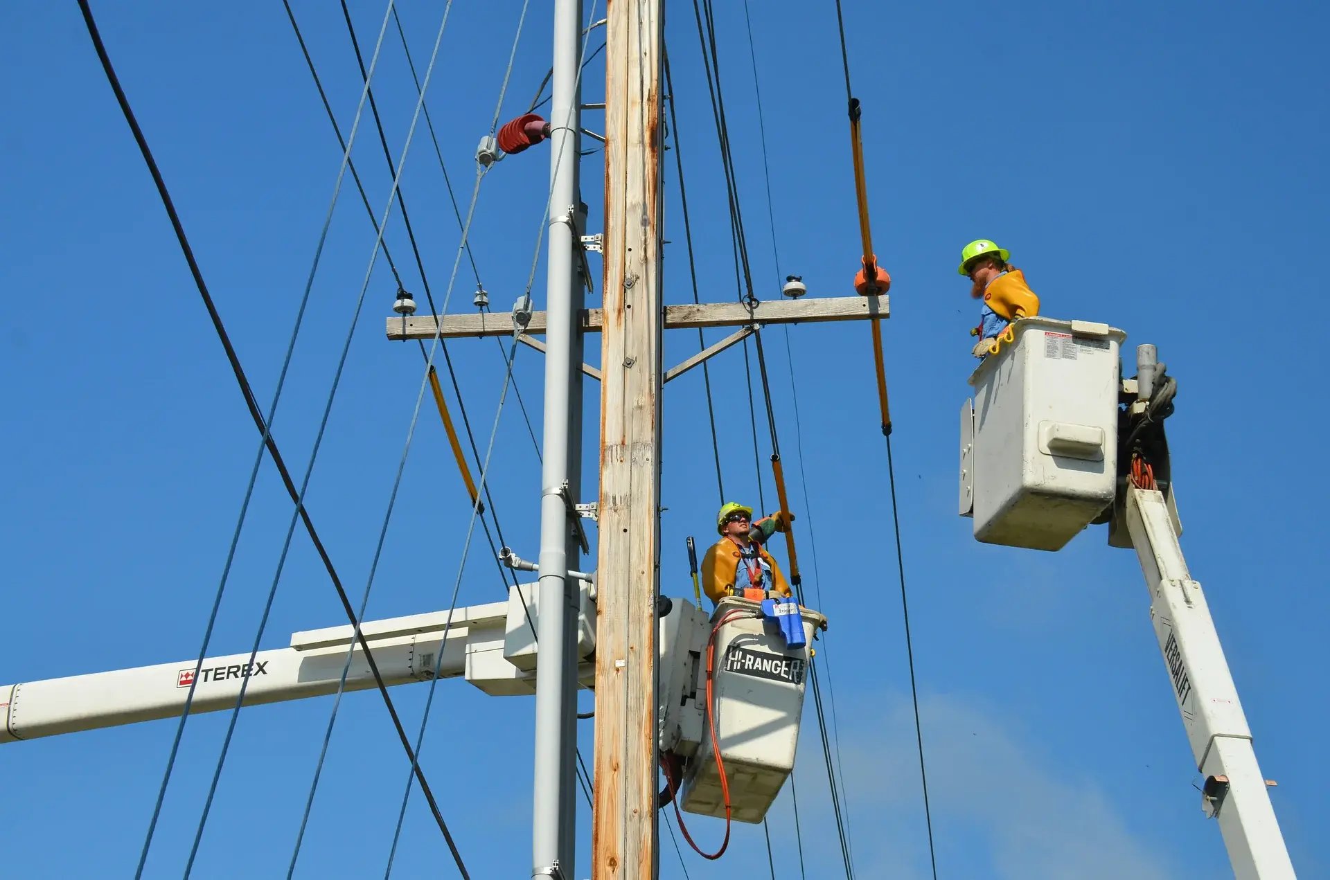 Two utility linemen in bucket trucks repairing overhead power lines on a wooden distribution pole.