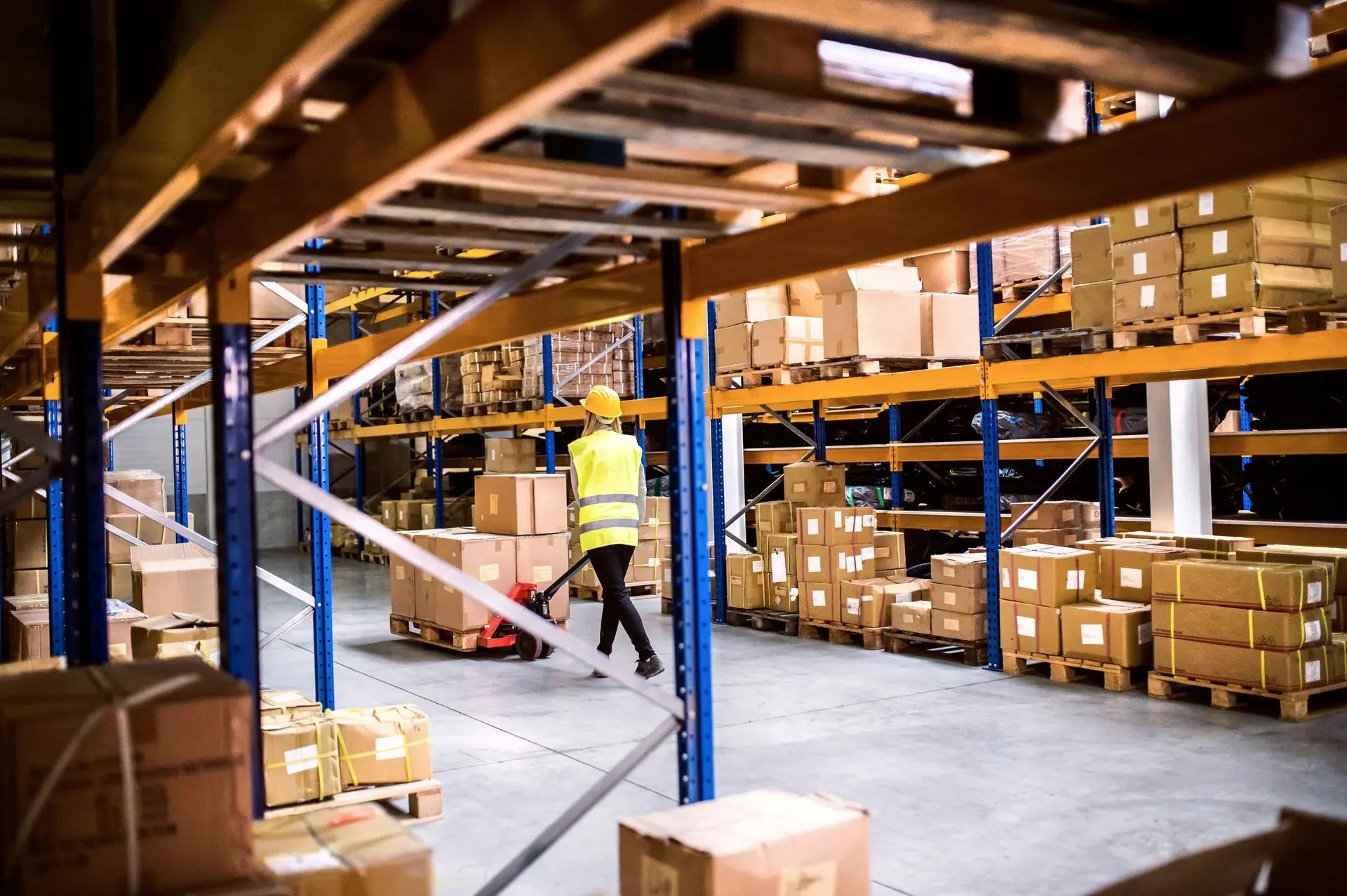 Warehouse worker in a yellow safety vest moving a loaded pallet jack through a distribution center.
