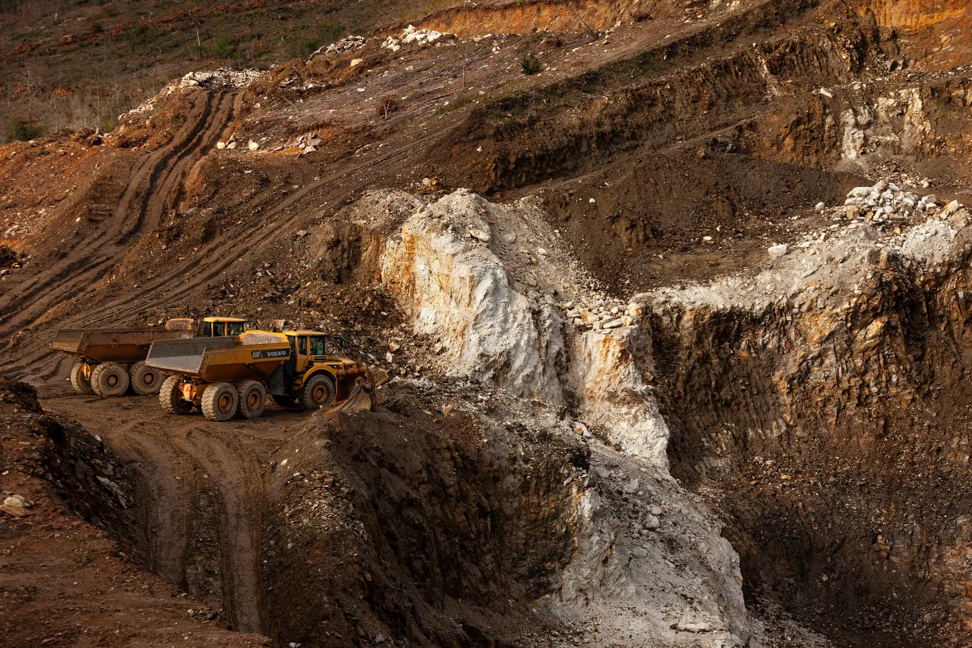 Two articulated haul trucks parked on a switchback road in an open-pit mining quarry at golden hour.