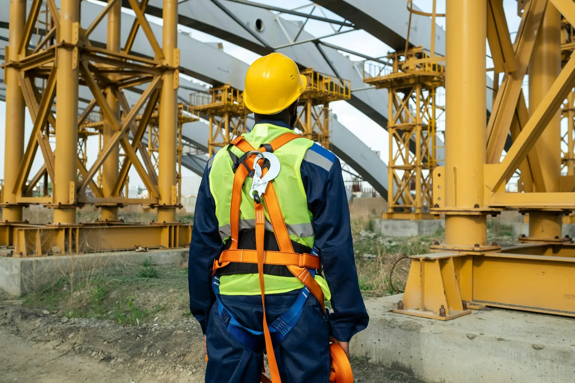 Construction worker in a hard hat, safety vest, and fall-protection harness facing a large yellow steel structure under construction.