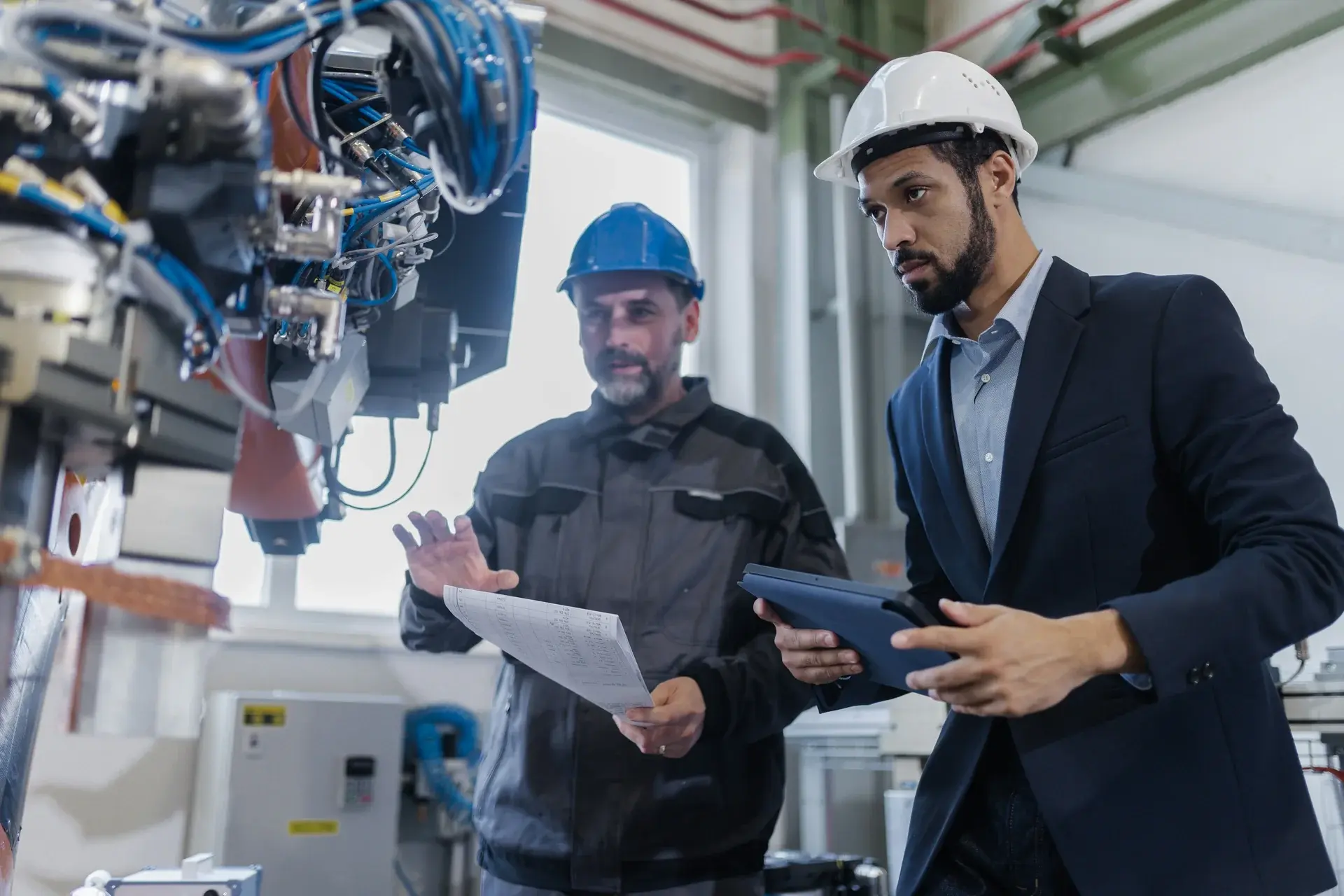 Engineer in a suit and technician in a hard hat reviewing data on a tablet next to industrial robotics equipment.