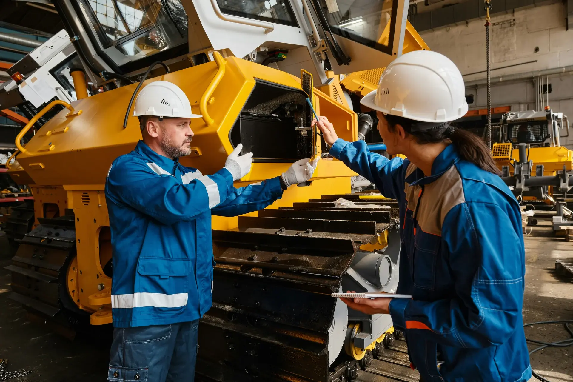 Two engineers in hard hats inspecting a yellow bulldozer in a heavy equipment manufacturing facility.