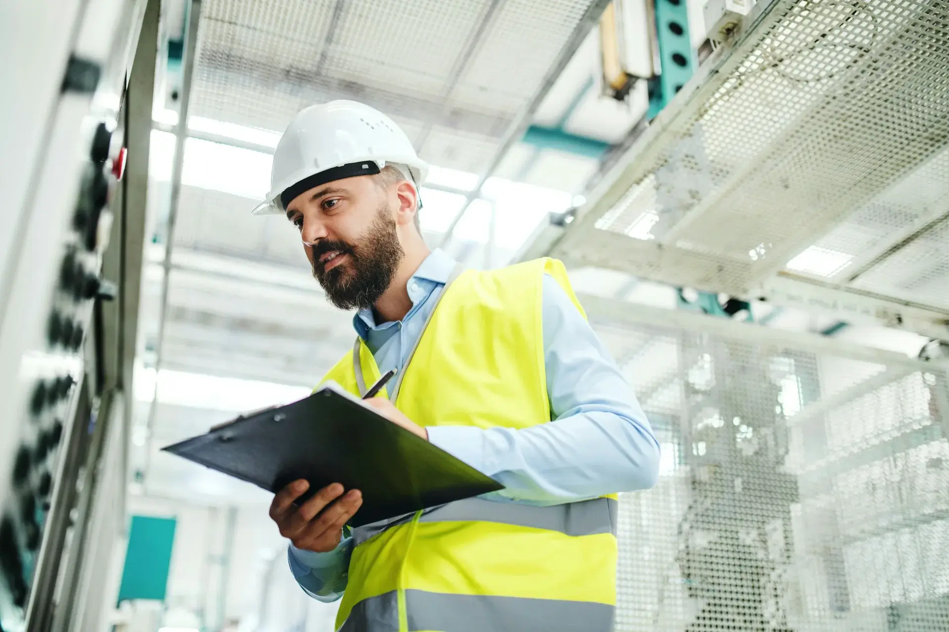 Industrial engineer in a hard hat and safety vest taking notes during an equipment inspection.