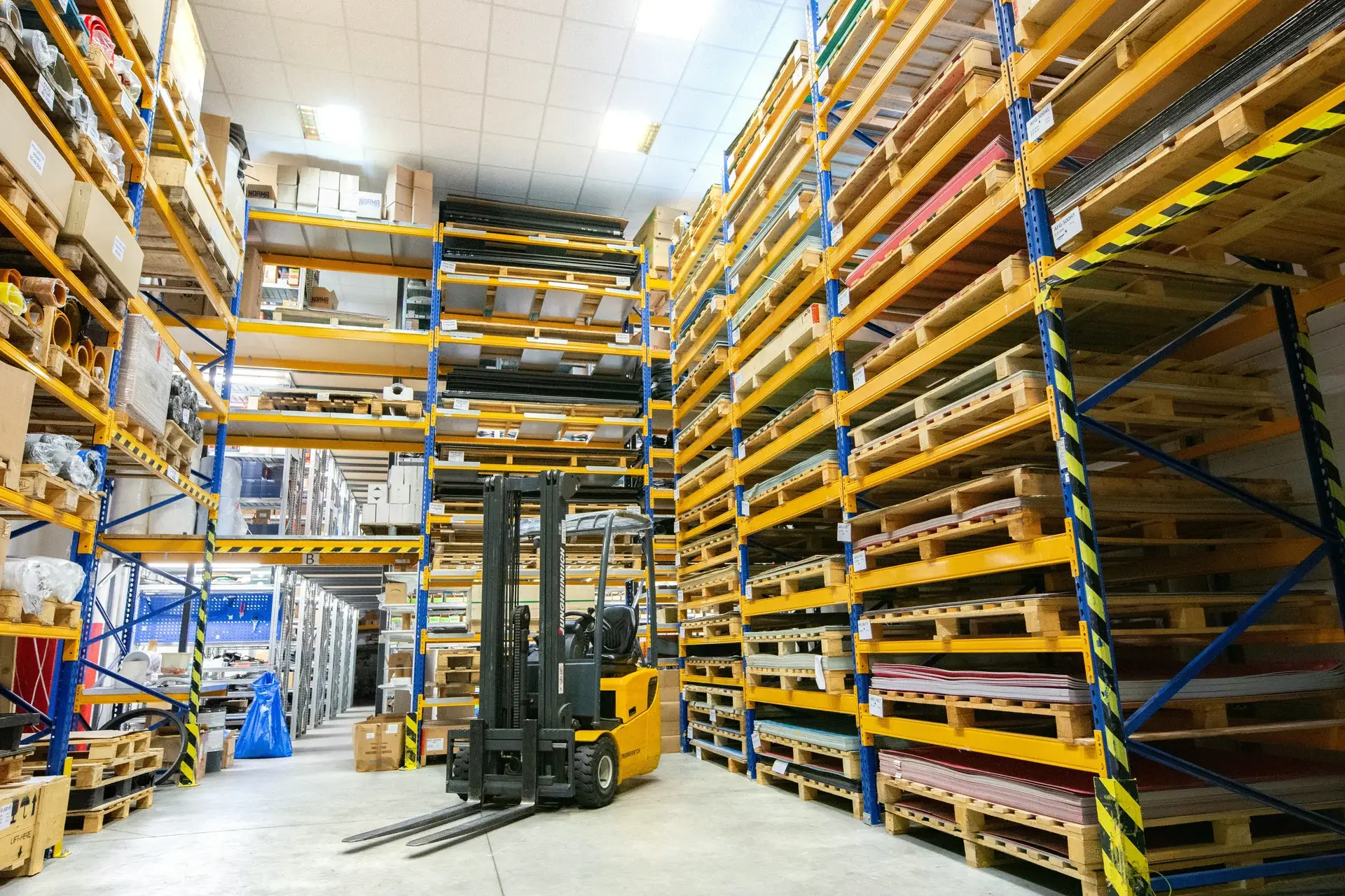 Industrial warehouse aisle with a parked forklift and tall pallet racks holding stacks of materials and sheet stock.