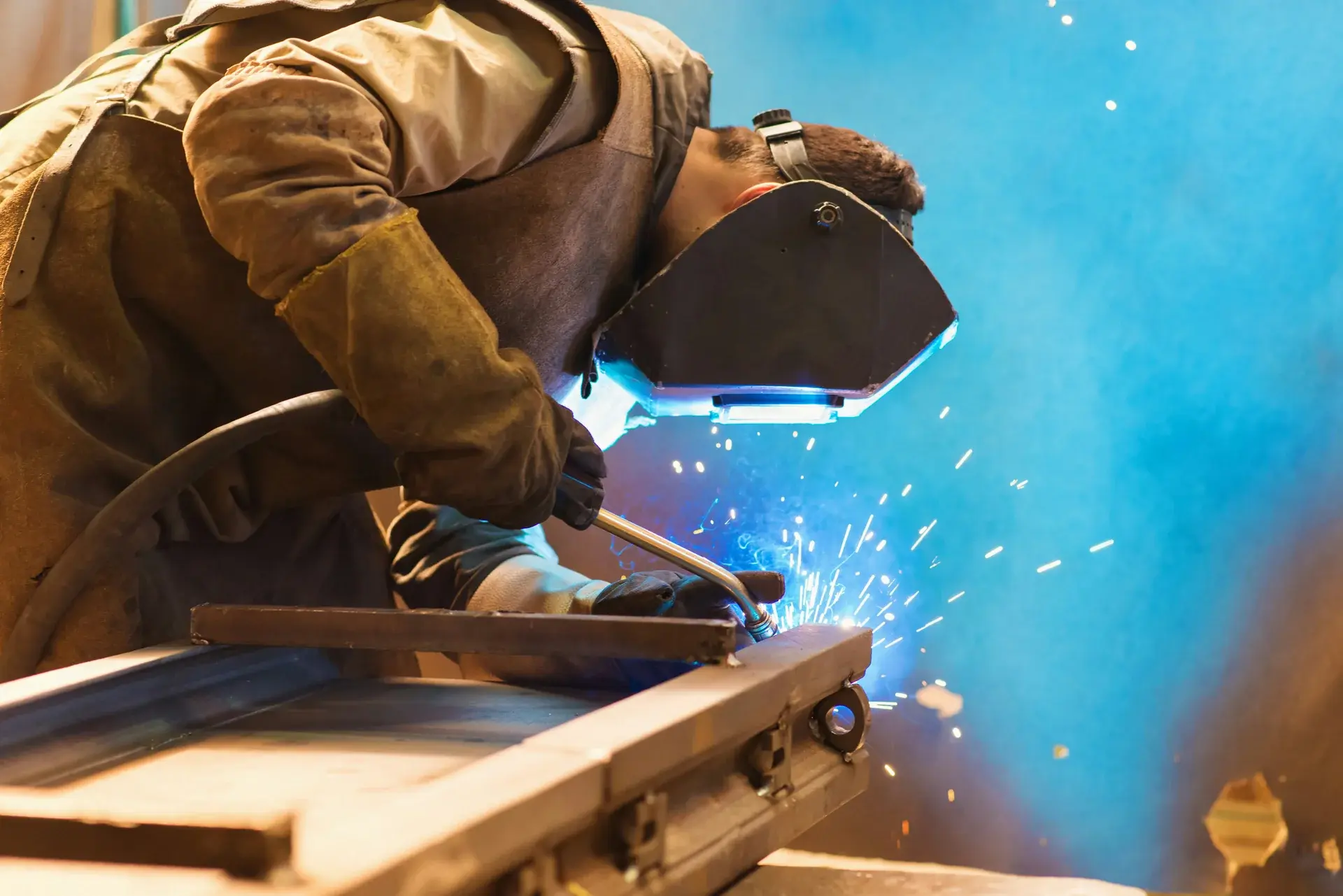 Industrial welder in protective gear MIG welding a steel workpiece with bright sparks and arc light.