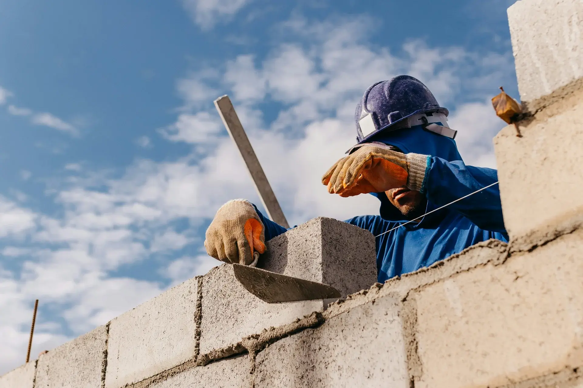 Construction mason in a hard hat laying a concrete block wall with mortar against a blue sky.