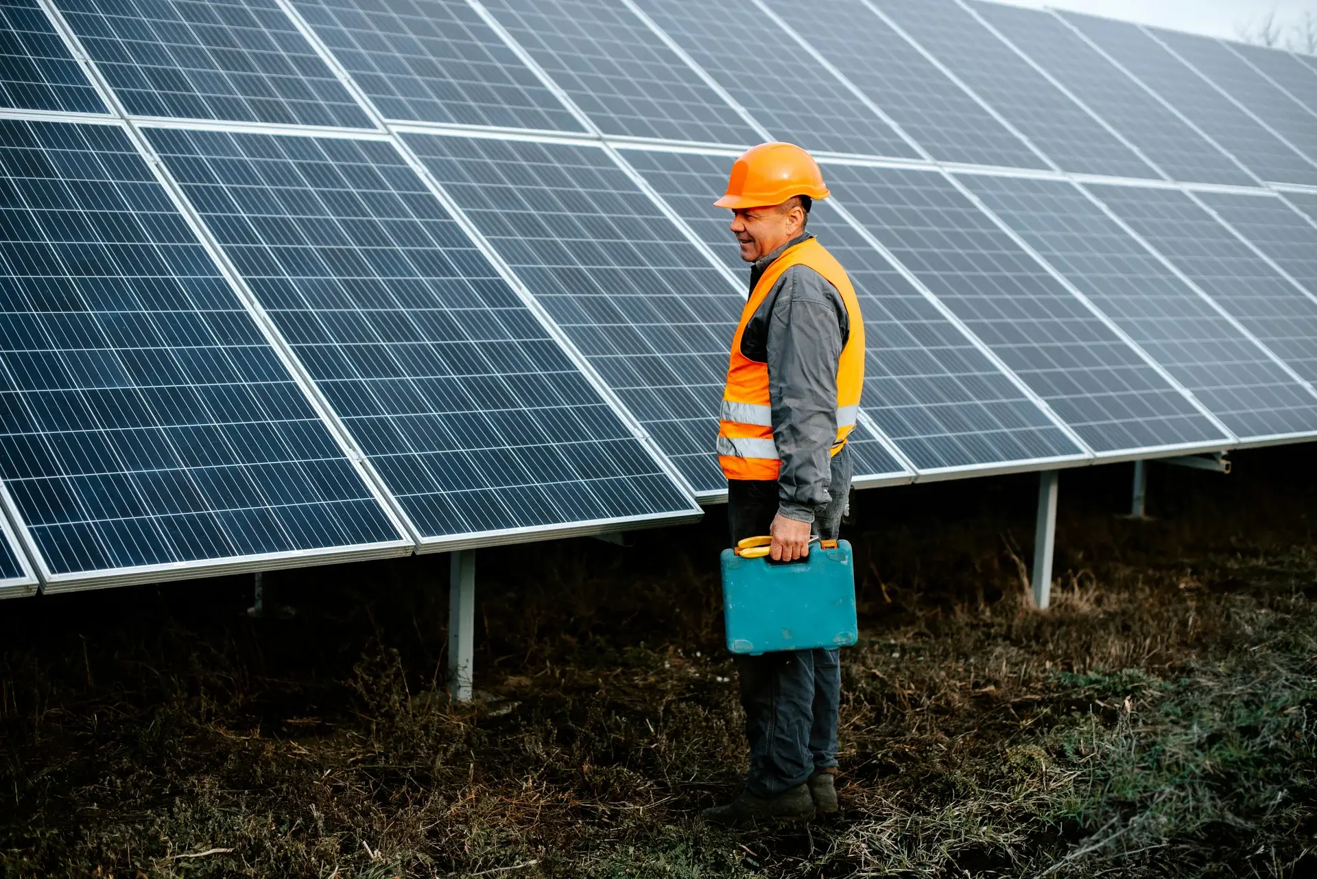 Solar technician in orange safety vest and hard hat carrying a toolbox beside a ground-mounted photovoltaic panel array.