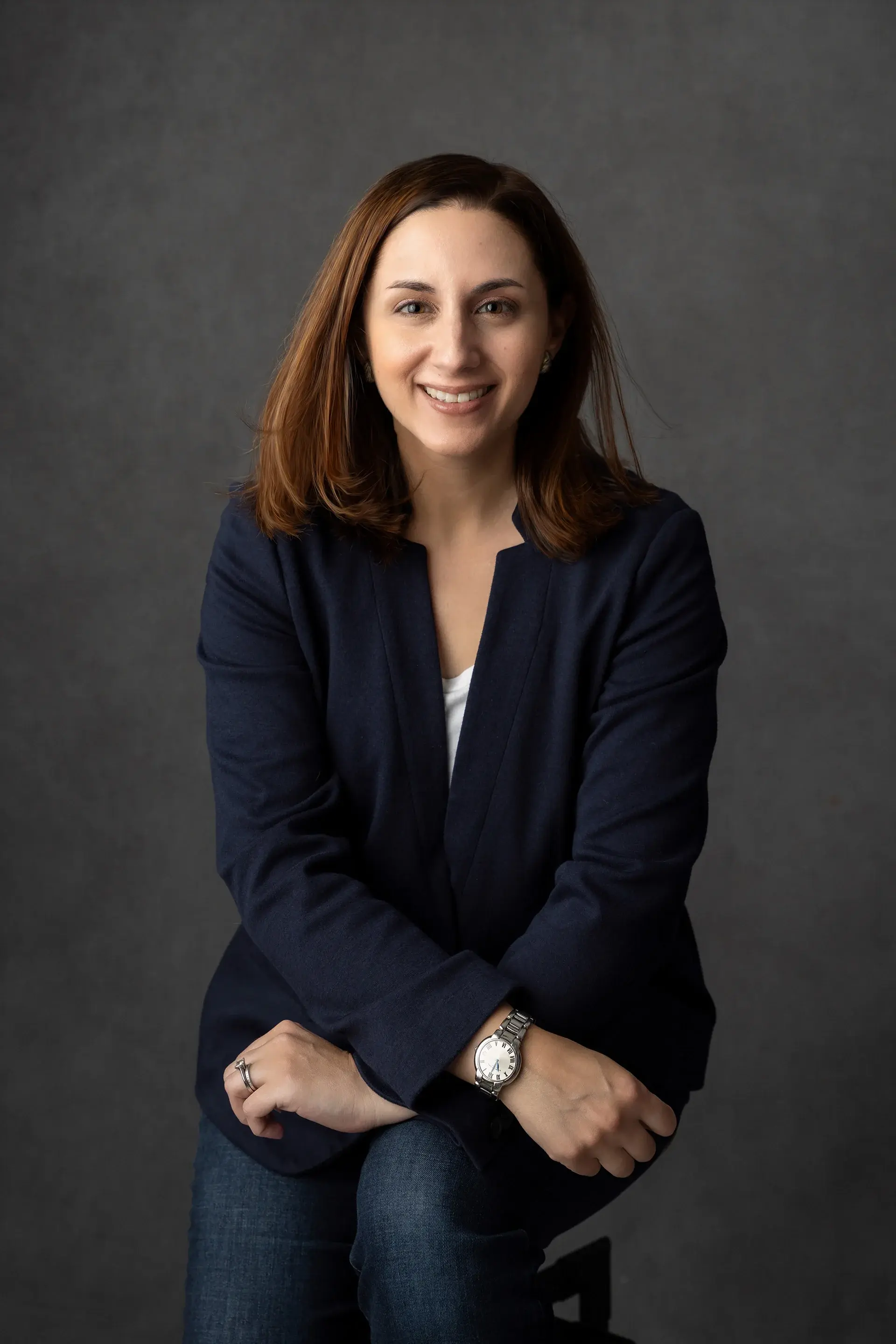 Professional headshot of a smiling team member in a navy blazer against a dark gray studio background.