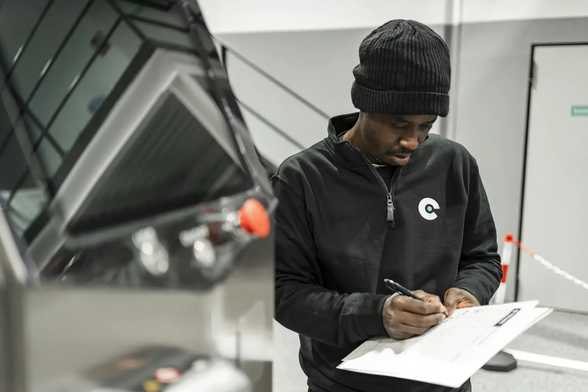 Technician in a black sweatshirt completing a paperwork checklist beside industrial equipment.
