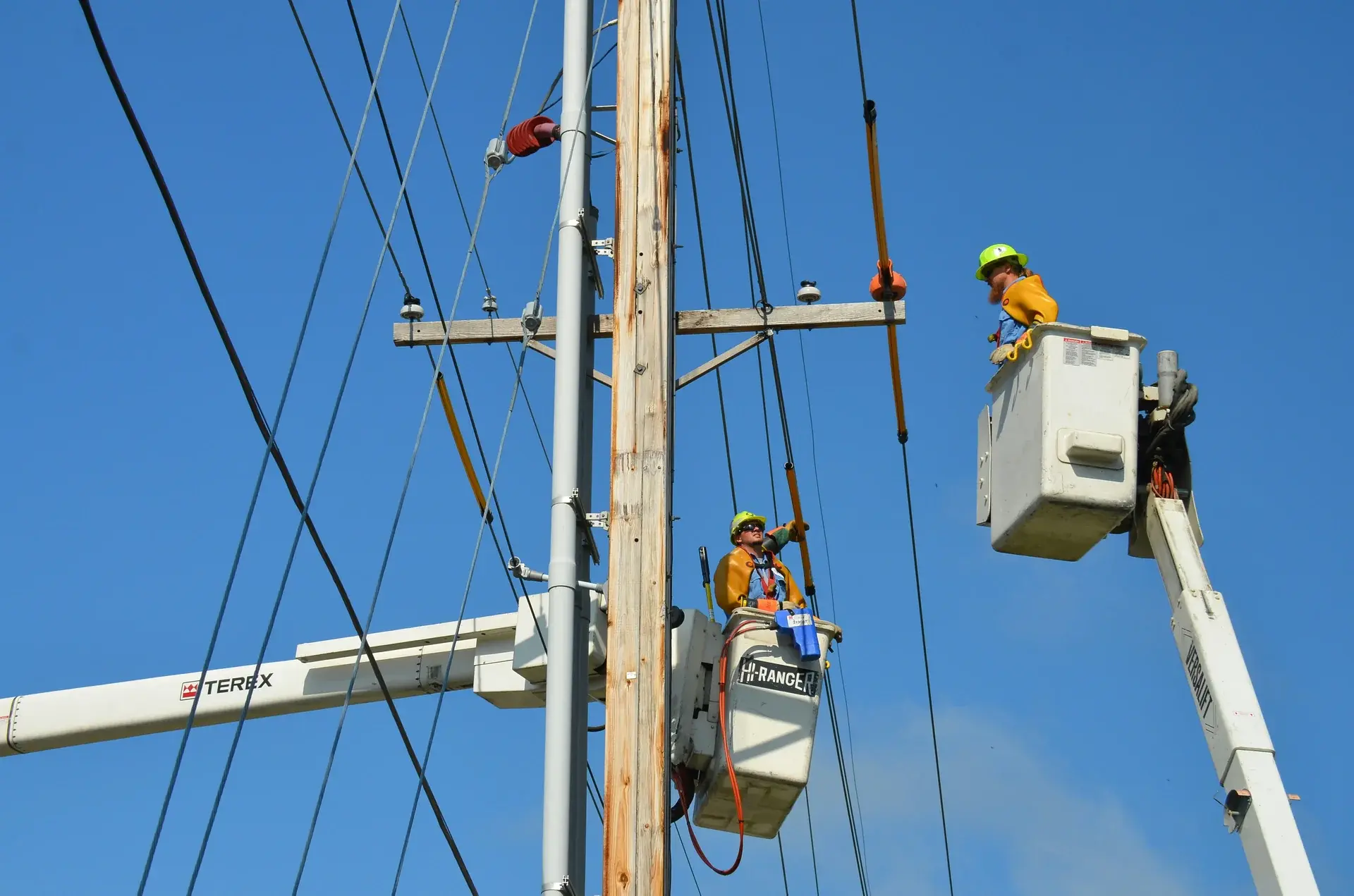 Two utility linemen in bucket trucks repairing overhead power lines on a wooden distribution pole.