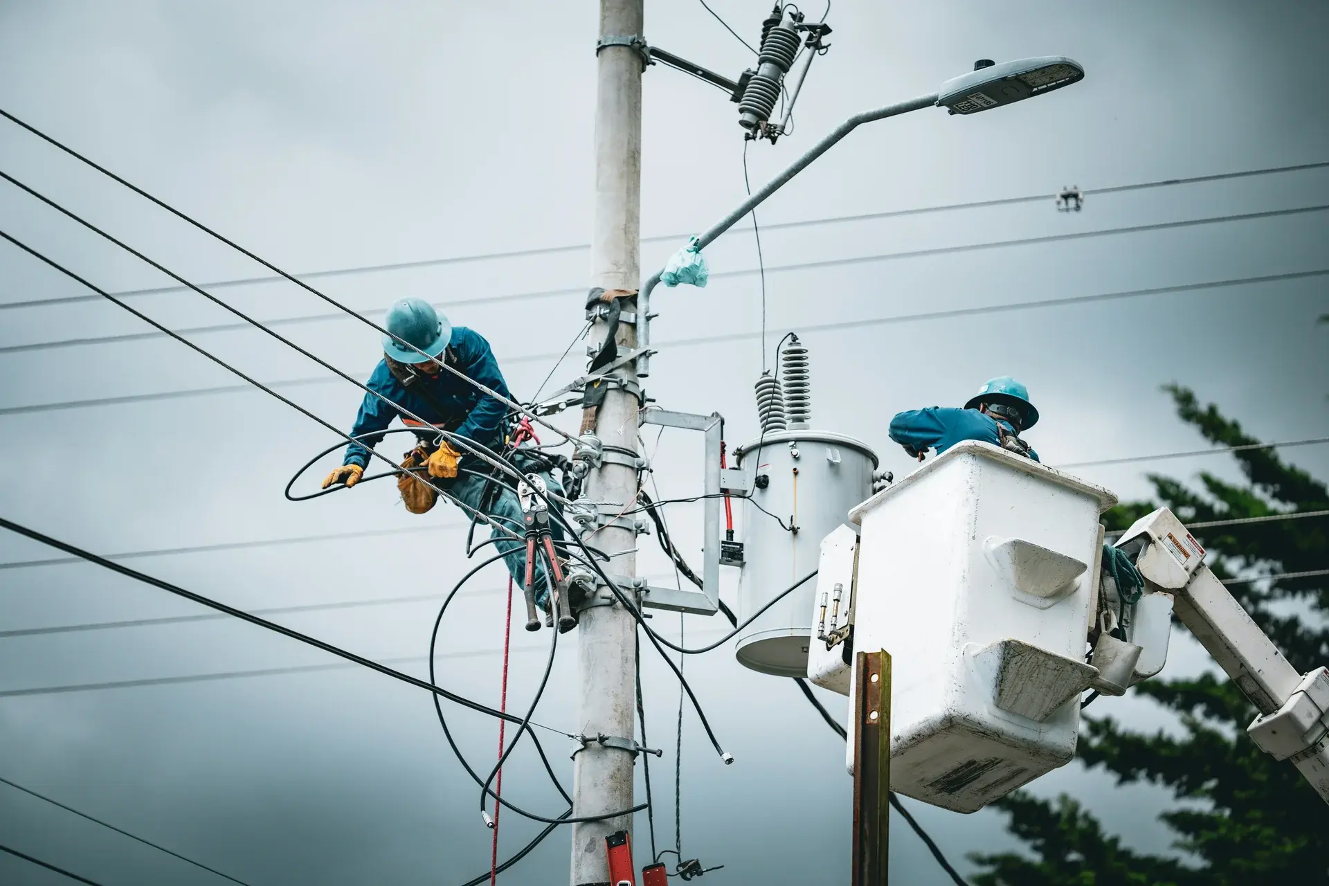 Two utility linemen repairing overhead power lines and a pole-mounted transformer against an overcast sky.