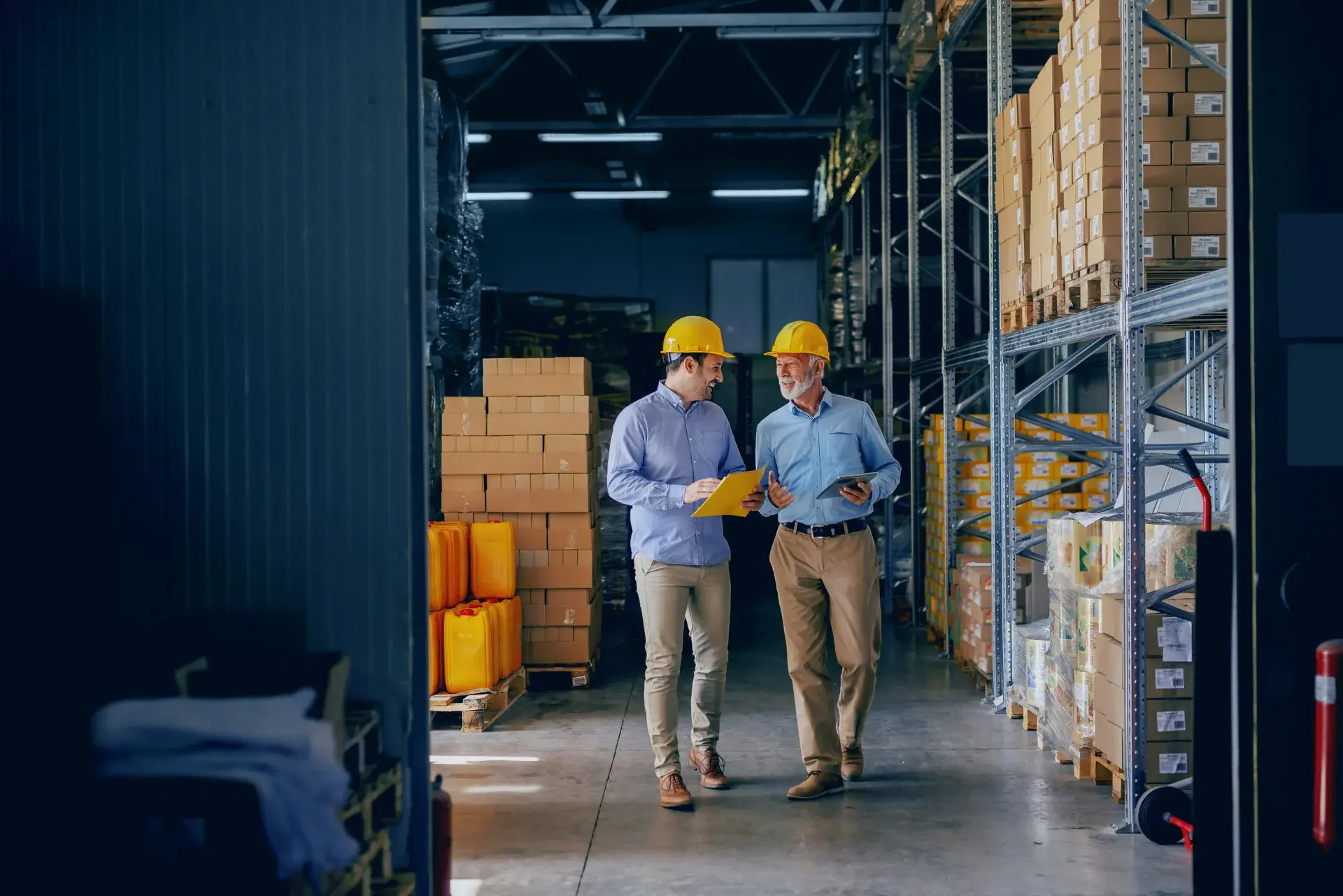 Two warehouse managers in hard hats reviewing inventory and clipboards inside an industrial storage facility.